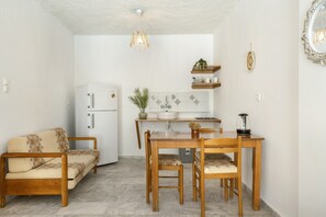Un espace de vie compact comprenant un canapé, une table à manger, des chaises, un réfrigérateur, un coin cuisine avec un évier et un plan de travail, ainsi qu’un luminaire suspendu.