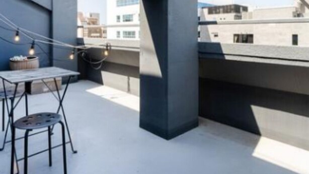 A rooftop terrace with a table and chairs, string lights, and a view of surrounding buildings.