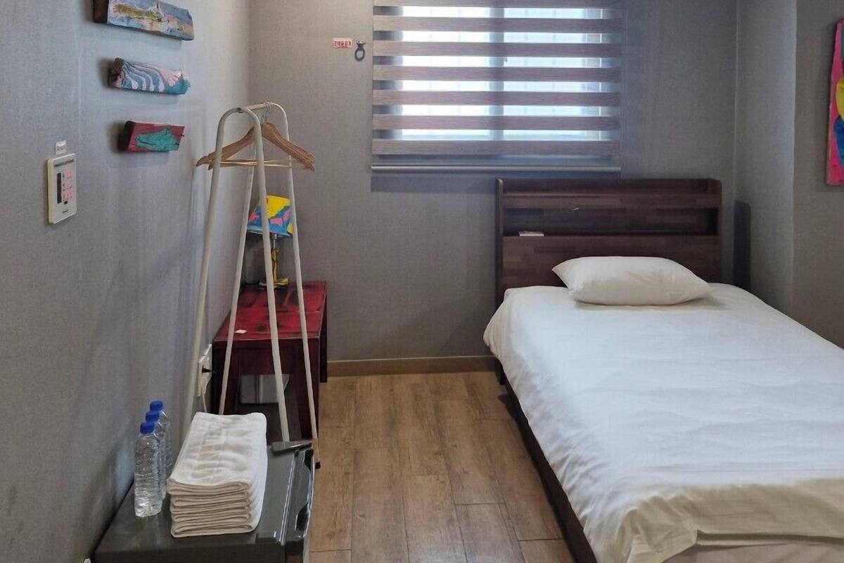 Une petite pièce bien éclairée, avec un lit, un plancher en bois, une fenêtre avec des stores et un radiateur fixé au mur.