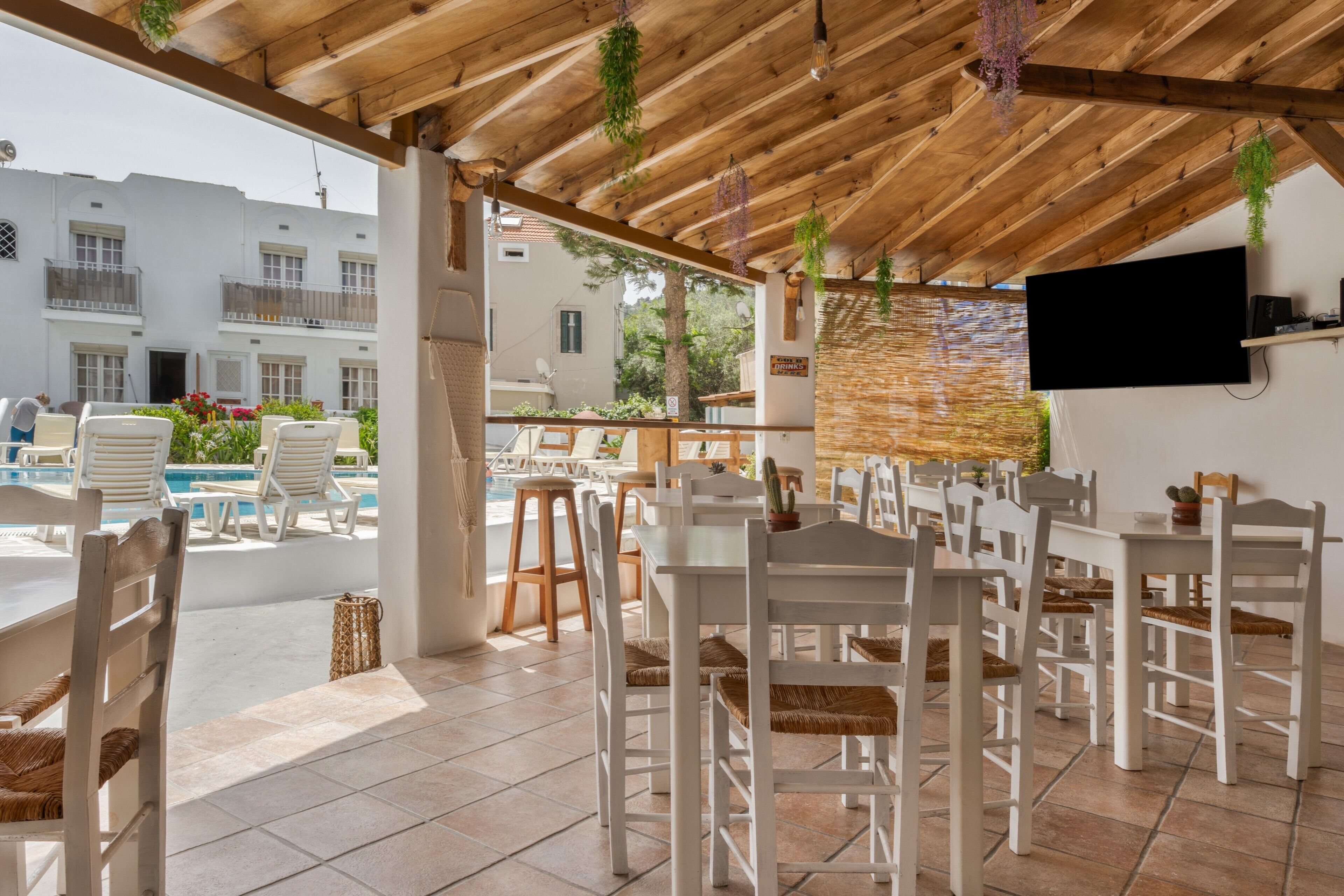 A covered outdoor seating area with white tables and chairs, a pool, and a TV mounted on the wall.