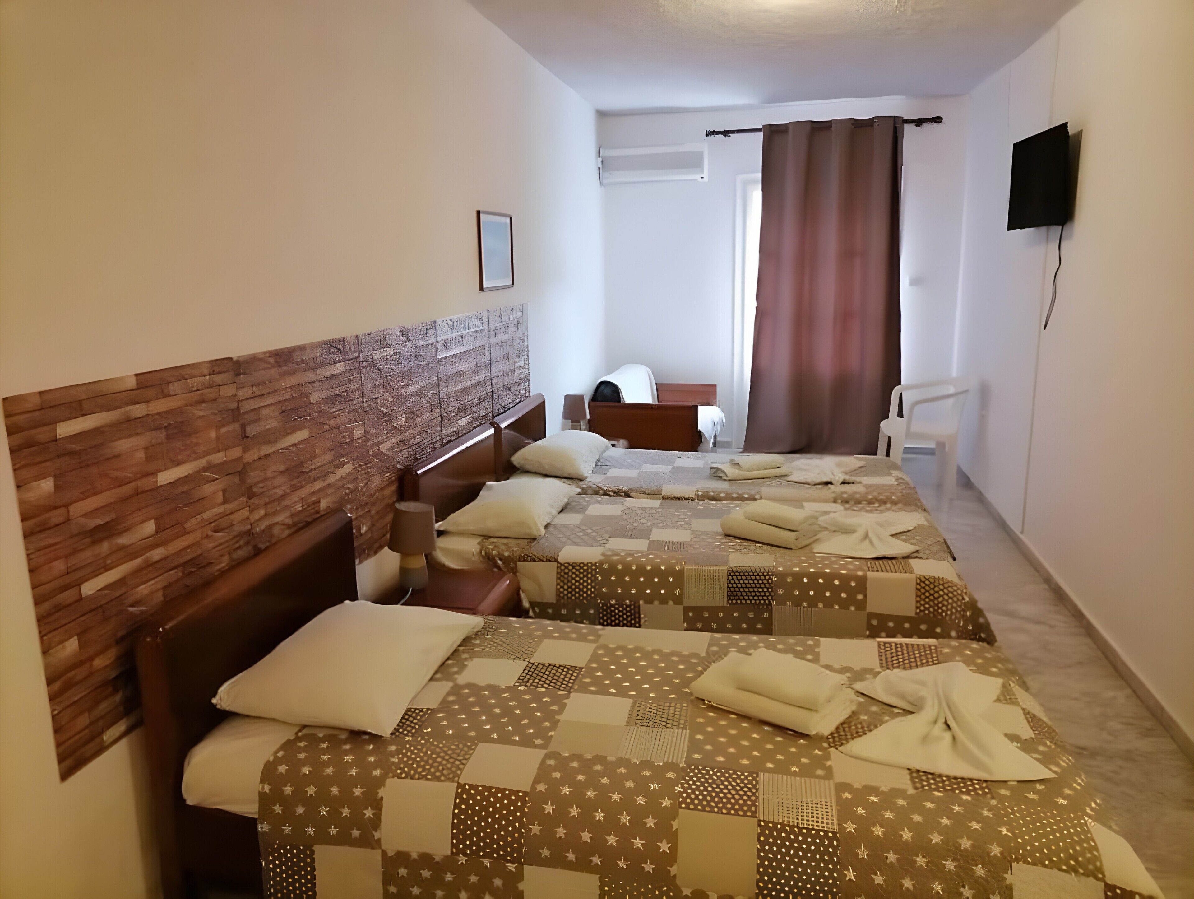 Ein Hotelzimmer mit zwei Betten, einem Holz-Kopfteil, einem Flachbildfernseher, einem Fenster mit Vorhängen und einem weißen Stuhl.