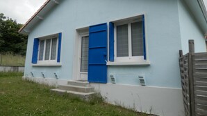 Una casa de dos pisos con fachada azul claro, techo marrón y dos ventanas con contraventanas azules.