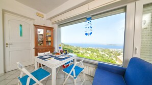 Quarto com mesa de jantar, cadeiras, um sofá, vista para o mar e uma janela com um adorno pendurado.