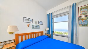 Uma cama com colcha azul, cabeceira de madeira, cadeira de vime, mesa de cabeceira com abajur e quadros na parede.