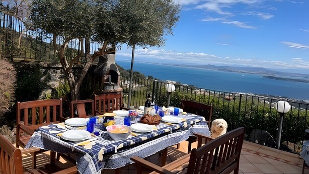 Área de refeições ao ar livre com mesa posta para refeição, com vista panorâmica para o mar e a terra distante.