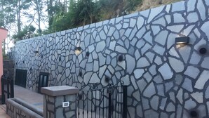 Mural de pedra com portão, plantas em vasos e luminárias externas.