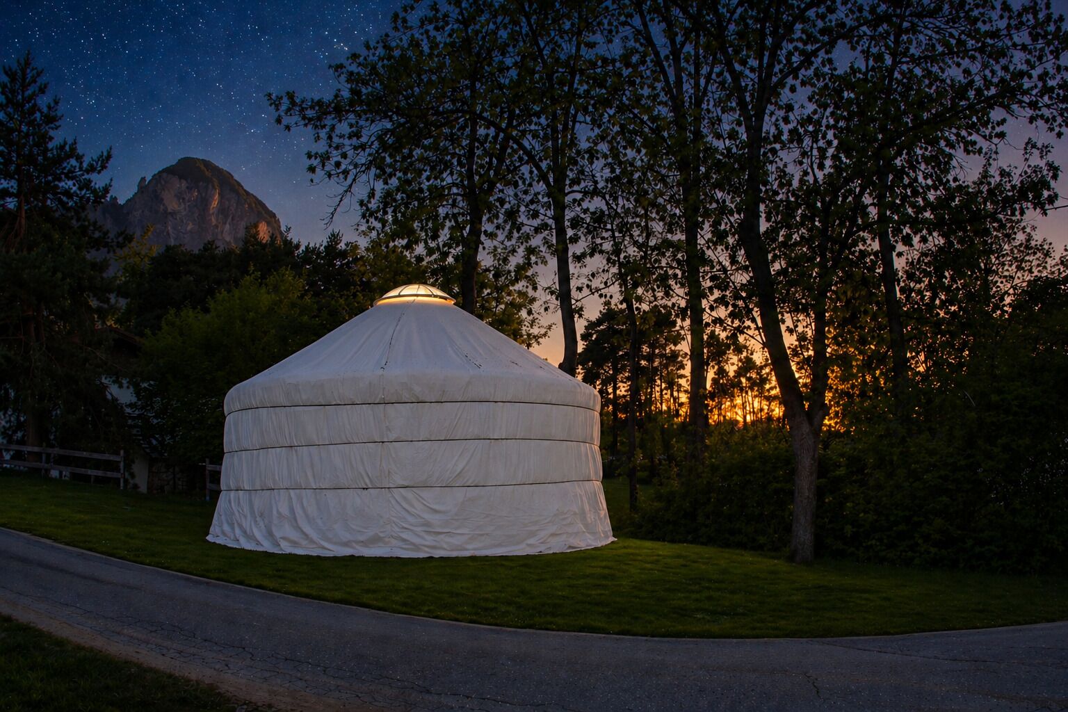 Una yurta con techo cónico, rodeada de árboles y una montaña al fondo.