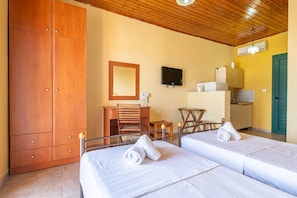 A hotel room with two beds, a wooden wardrobe, a desk with a chair, a television, and a kitchenette.