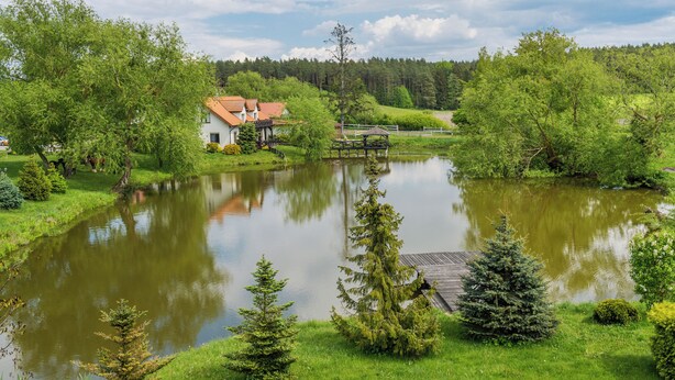 A serene lakeside setting with a wooden bridge, a small building with a red roof, and lush greenery. - Willa Sun & Snow Zasypane Mazury (gmina Malkinia Gorna)