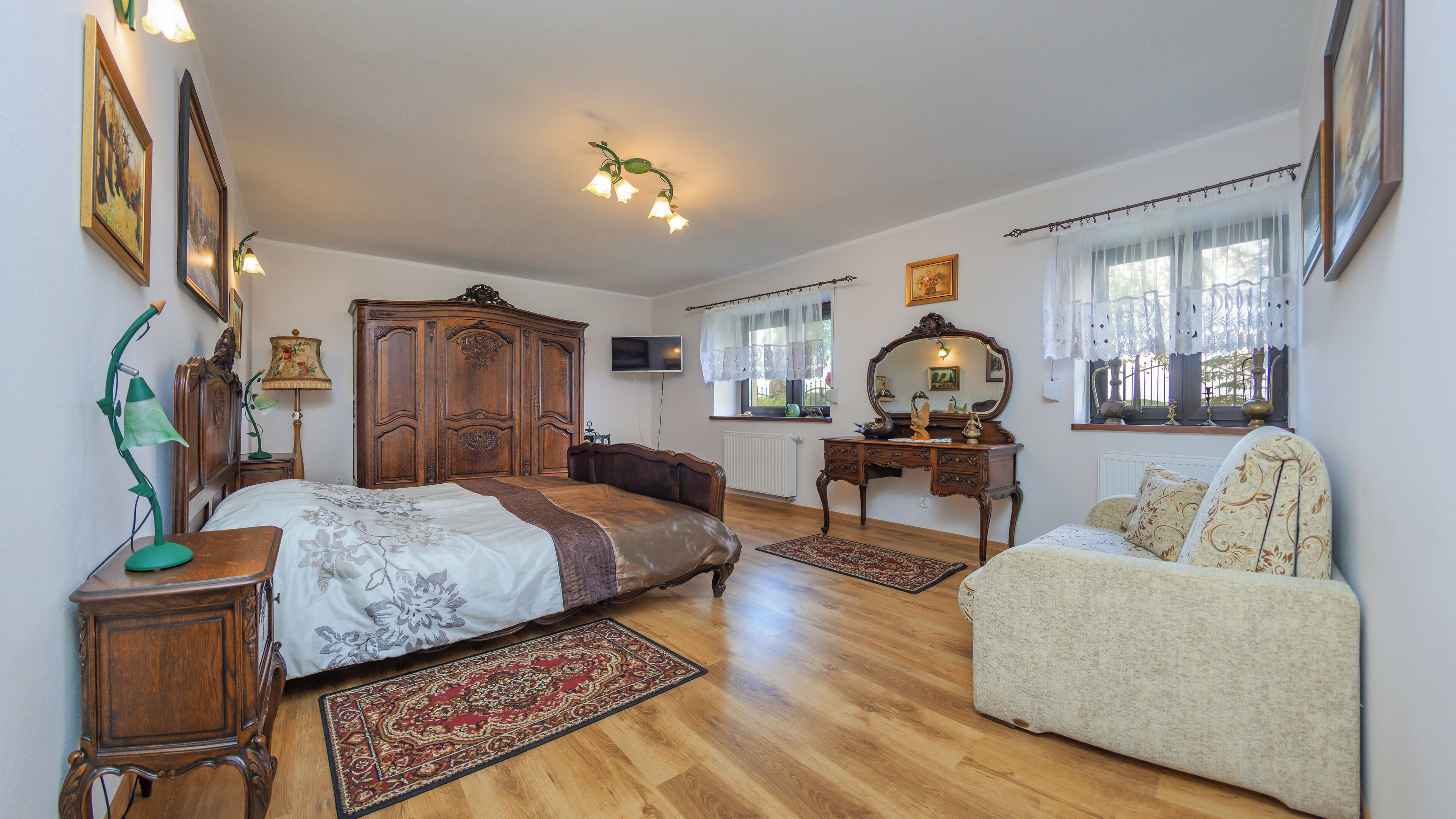 A spacious bedroom with a large bed, a wooden wardrobe, a desk, a chair, and a television on the wall.