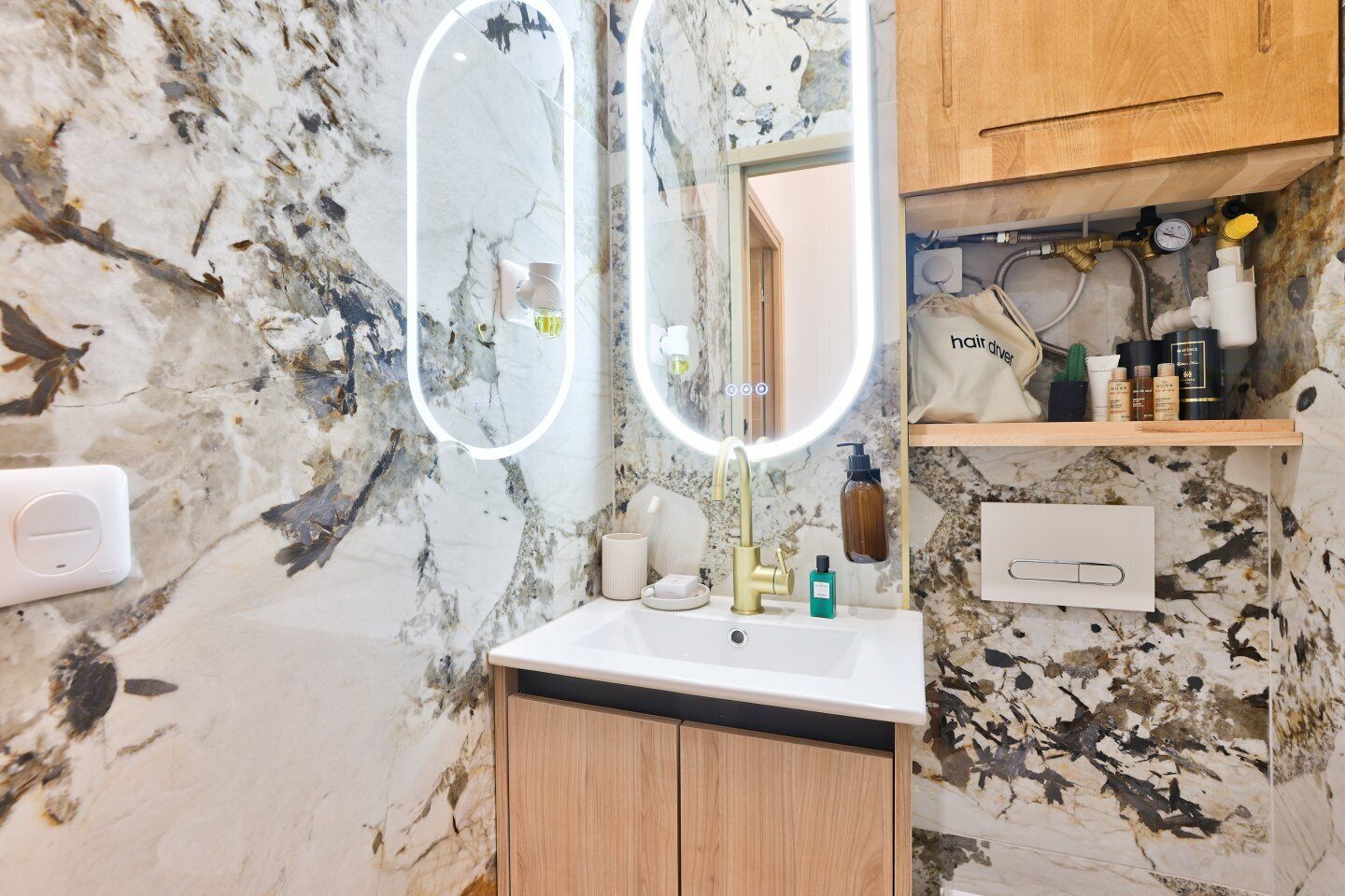 A bathroom with a marble wall, a sink with a gold faucet, and a mirror with a circular light.
