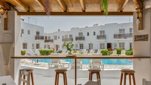 A poolside bar with white chairs and stools, a pool, and white buildings with balconies in the background.