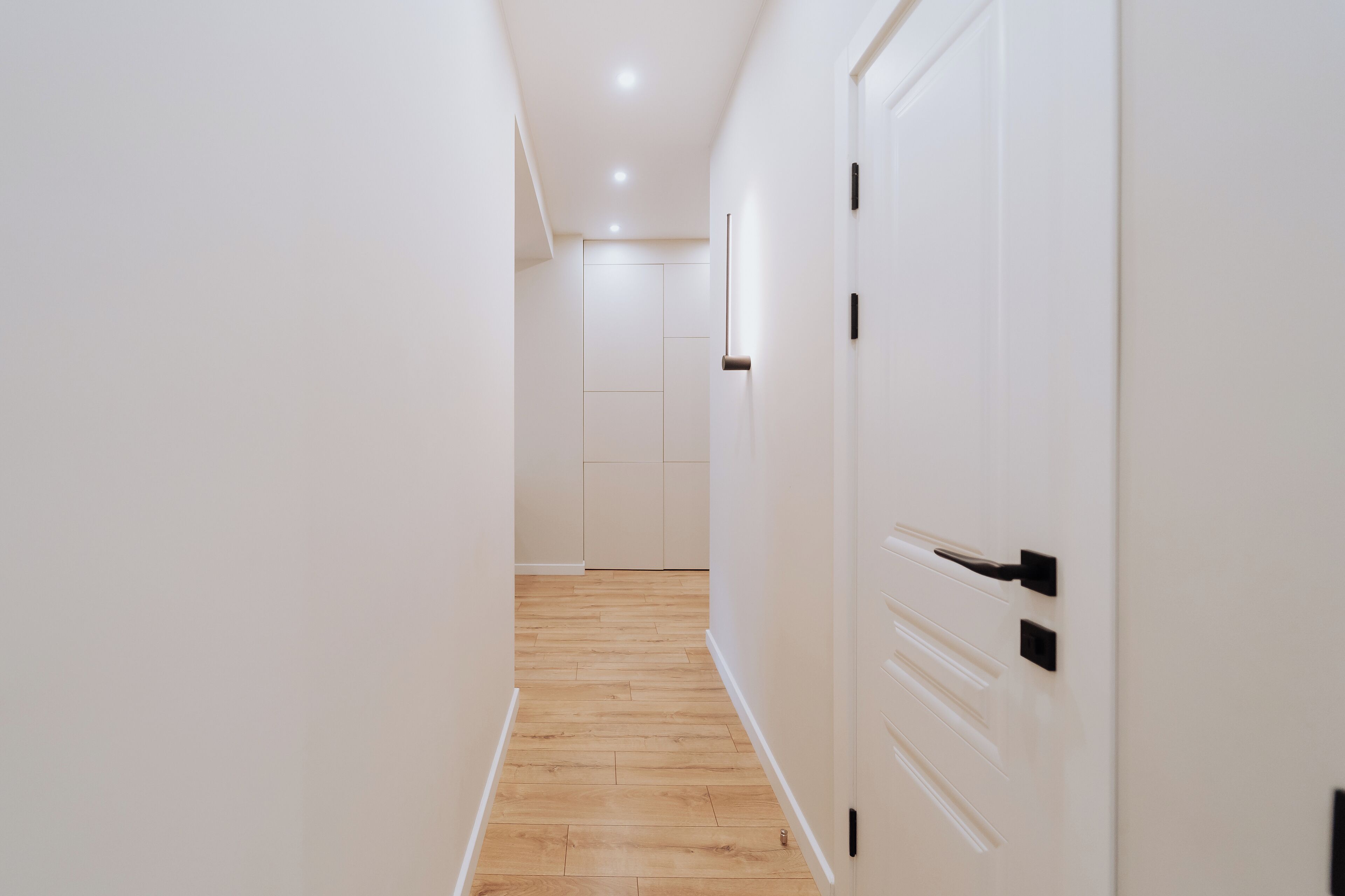 A narrow hallway with wooden flooring and white walls. There are two white doors with black handles on the right side.