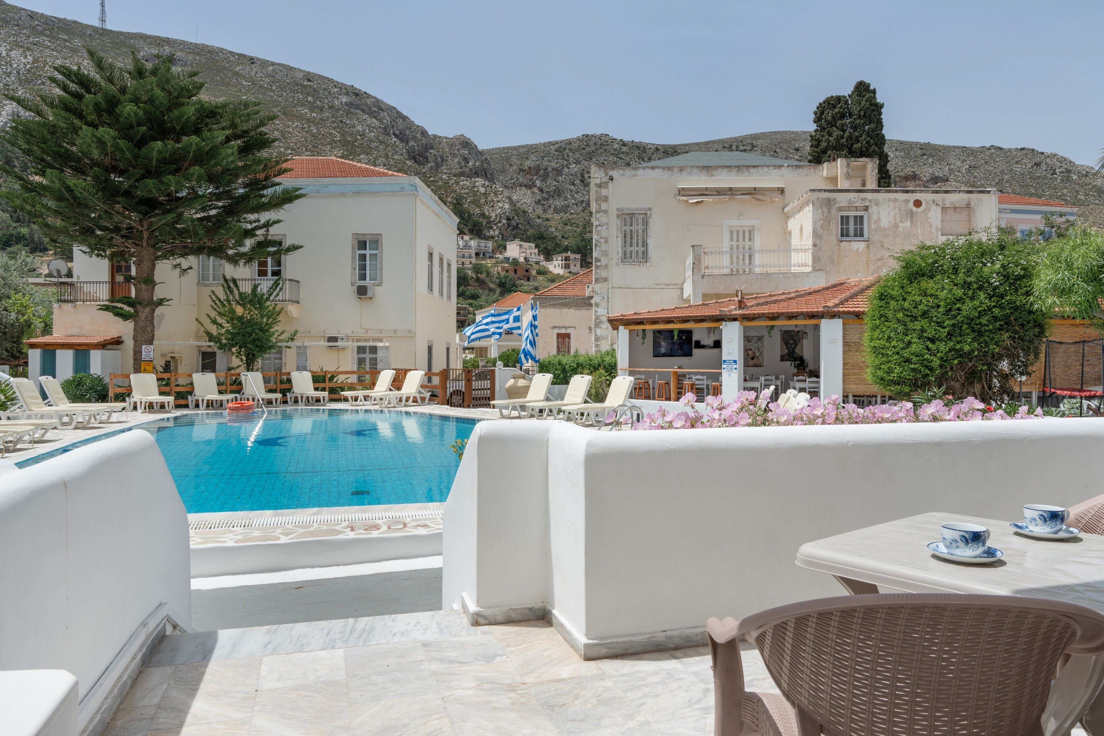 Uma área de piscina com espreguiçadeiras e uma mesa, cercada por edifícios com varandas e um cenário montanhoso.