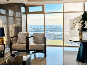 A spacious room with a large window offering a view of a coastal landscape. There are two armchairs and a small table with a lamp.