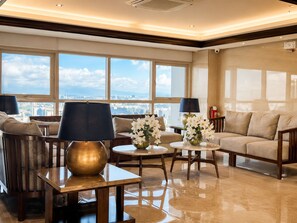A spacious lounge area with a sofa set, two side tables, and a large window offering a city view.