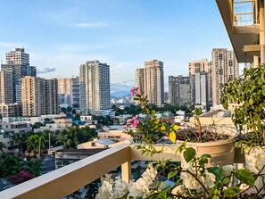 A cityscape with high-rise buildings, a balcony with potted plants, and a clear sky.