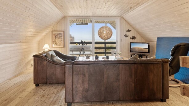A cozy attic room with a wooden ceiling, a large window with a view, a leather sofa, a television, and a blue chair. - "Naina" - 200m from the sea by Interhome (Grenaa)