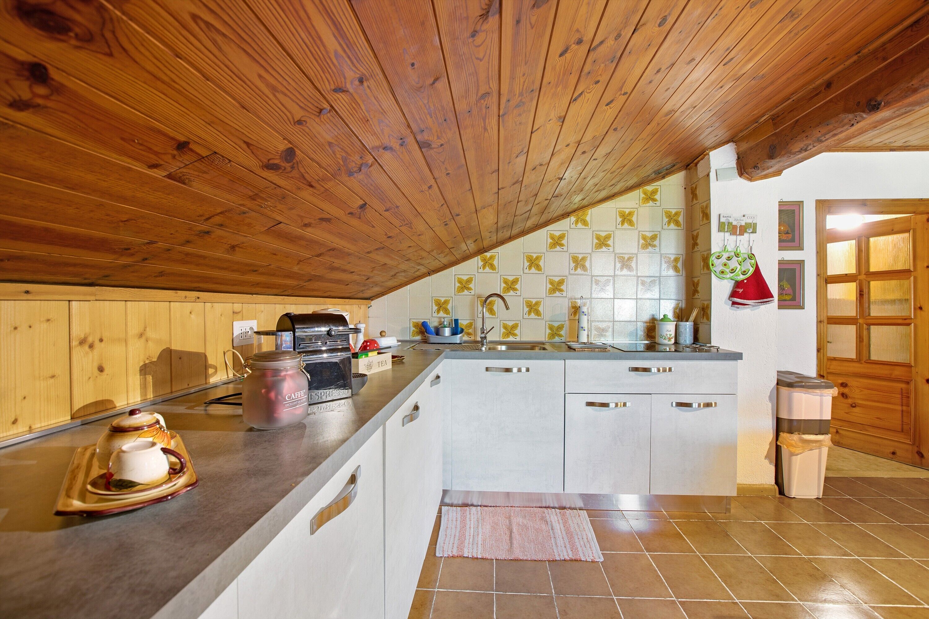 Une cuisine avec un plafond en bois, des armoires blanches, un pare-feu en carrelage et un plan de travail sur lequel sont disposés divers objets.