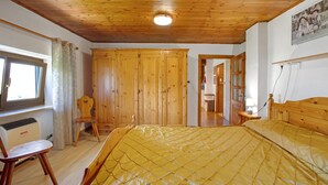 Ein Schlafzimmer mit Holzbalkendecke, einem Bett mit gelber Steppdecke, Holzschränken, einem Fenster mit Vorhängen und einem Heizkörper.