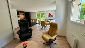 Uma sala de estar moderna com sofá, poltrona, mesa de jantar e cadeiras. Uma cozinha visível ao fundo.