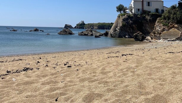 Paisagem costeira com praia arenosa, um edifício branco com chaminé e afloramentos rochosos ao longe.