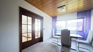 Ein Zimmer mit Holzboden, einer Schiebetür zum Balkon, einem Esstisch mit Stühlen und einem Fenster mit violetten Vorhängen.