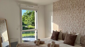 A bedroom with a large bed, a window with a view of greenery, a mirror, and patterned wallpaper.