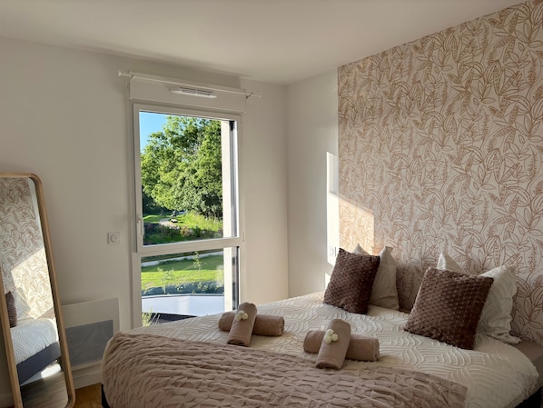 Une chambre à coucher avec un grand lit, une fenêtre donnant sur un paysage verdoyant, un miroir et un papier peint à motifs.
