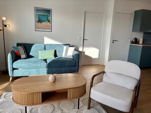 En stue med sofa, sofabord, stol, et maleri og et tekøkken.