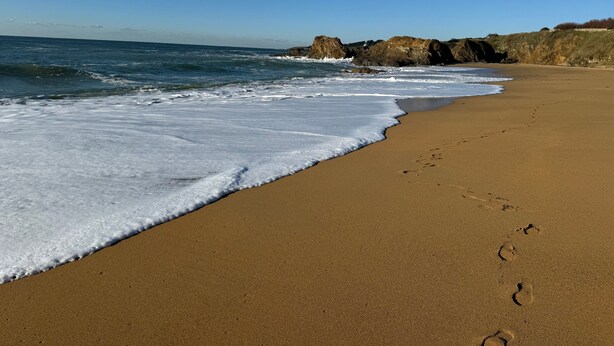 A sandy beach with footprints leading to the water, a clear sky, and rocky outcrops in the background.