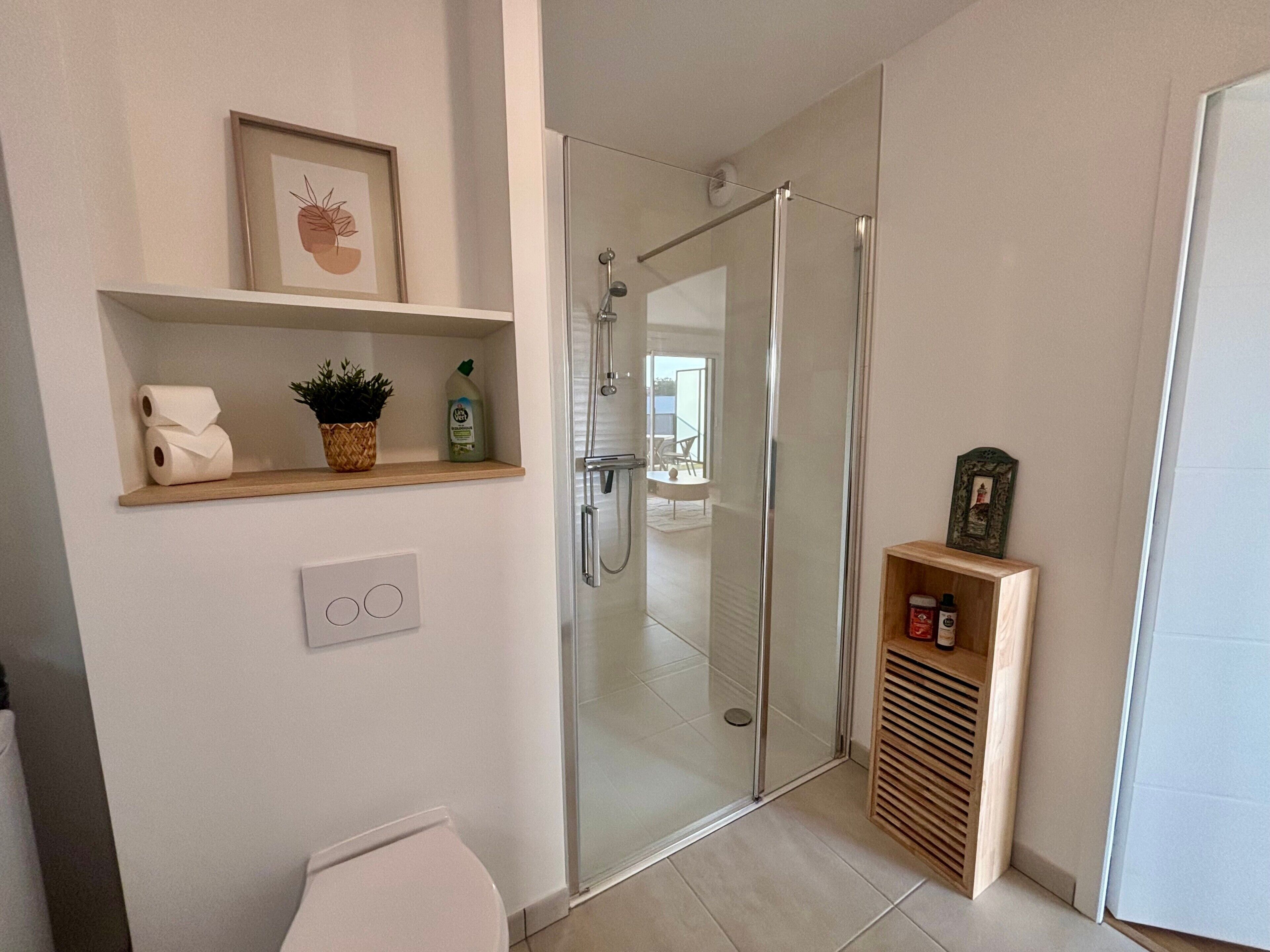 Une salle de bain moderne avec une douche à parois de verre, des toilettes, une étagère contenant des articles de toilette et un tableau encadré.