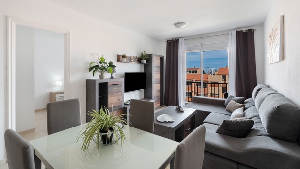 A modern living room with a dining area, a flat-screen TV, a sofa, and a balcony with a view of buildings. - Vacation 'Gabriela' with Shared Pool, Balcony and Wi-Fi (Candelaria)