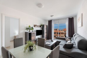 A modern living room with a dining area, a flat-screen TV, a sofa, and a balcony with a view of buildings.