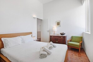 A hotel room with a bed, a wooden nightstand, a green chair, and a small table with a lamp.