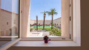 Une fenêtre donnant sur une cour intérieure agrémentée de palmiers, d’une piscine et d’un bâtiment orné d’arcades.