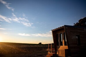 A modern wooden cabin with a staircase leading to the entrance, situated in a field with a clear sky.