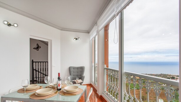 A dining area with a table set for four, a bottle of wine, and a view of the sea through large windows.
