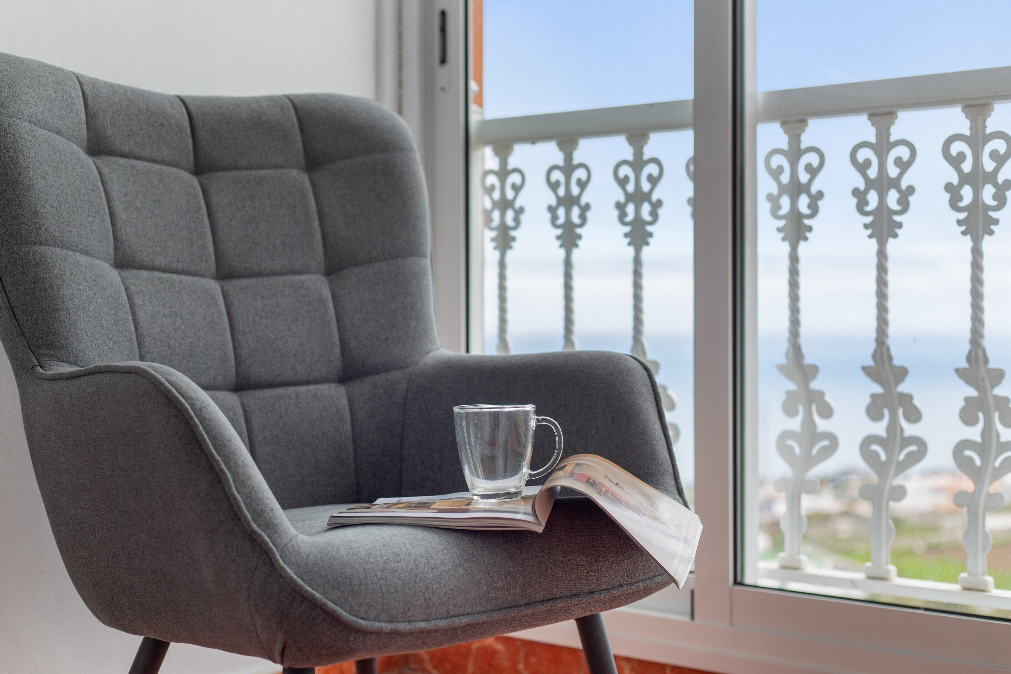 Un sillón tapizado gris con una taza de vidrio transparente y una revista, ubicado junto a una ventana con barandilla blanca.