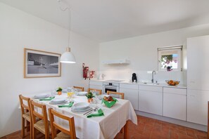 A dining area with a white table set for four, a kitchen with white cabinets, a white pendant light, and a framed picture on the wall.