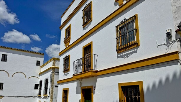 Un edificio blanco con detalles amarillos, múltiples ventanas con barandillas negras y una puerta verde.