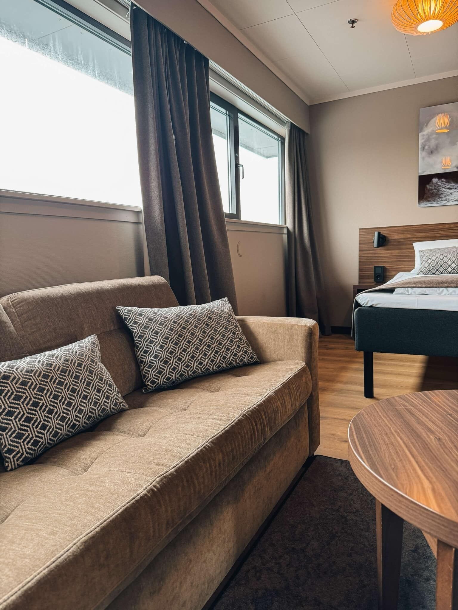 Une chambre d’hôtel moderne, équipée d’un canapé, d’un lit, d’une table ronde en bois et d’une grande fenêtre avec des rideaux.