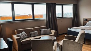 Una habitación de hotel moderna con sofá, dos sillones, una mesita, cama y ventanas amplias con vistas a la ciudad.