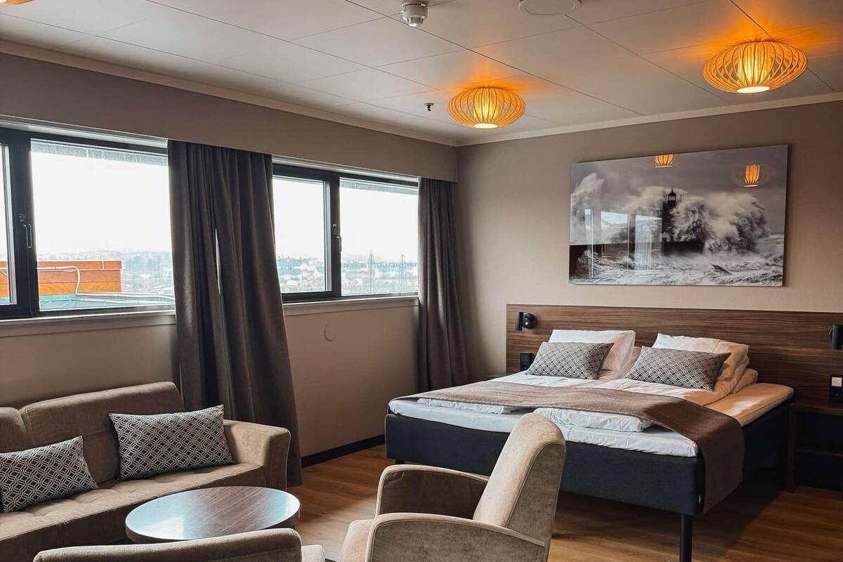 Ett modernt hotellrum med en stor säng, en soffa, ett runt bord och utsikt över staden genom stora fönster.