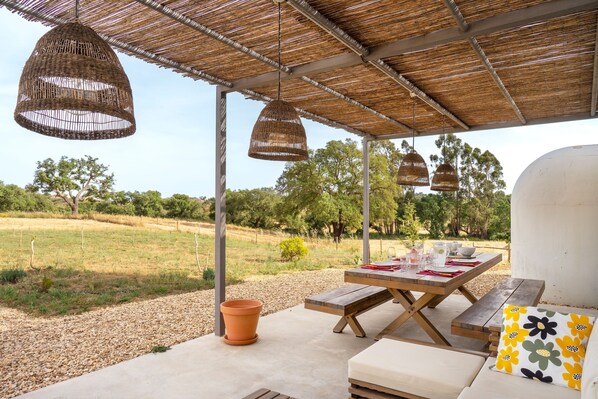 Une terrasse extérieure avec une table dressée pour le repas, un banc et des lampes en osier suspendues.