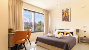 A modern bedroom with a large bed, a desk with a chair, and a window with a view of trees. - Nobia 8 (Athens)