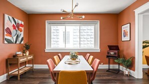 A dining area with a white table, orange chairs, a wooden sideboard, and a large window with a view of buildings. - Retro Indy stay-sleeps 10! Big backyard, outdoor games, BBQ, 5 min from Downtown (Indianapolis)