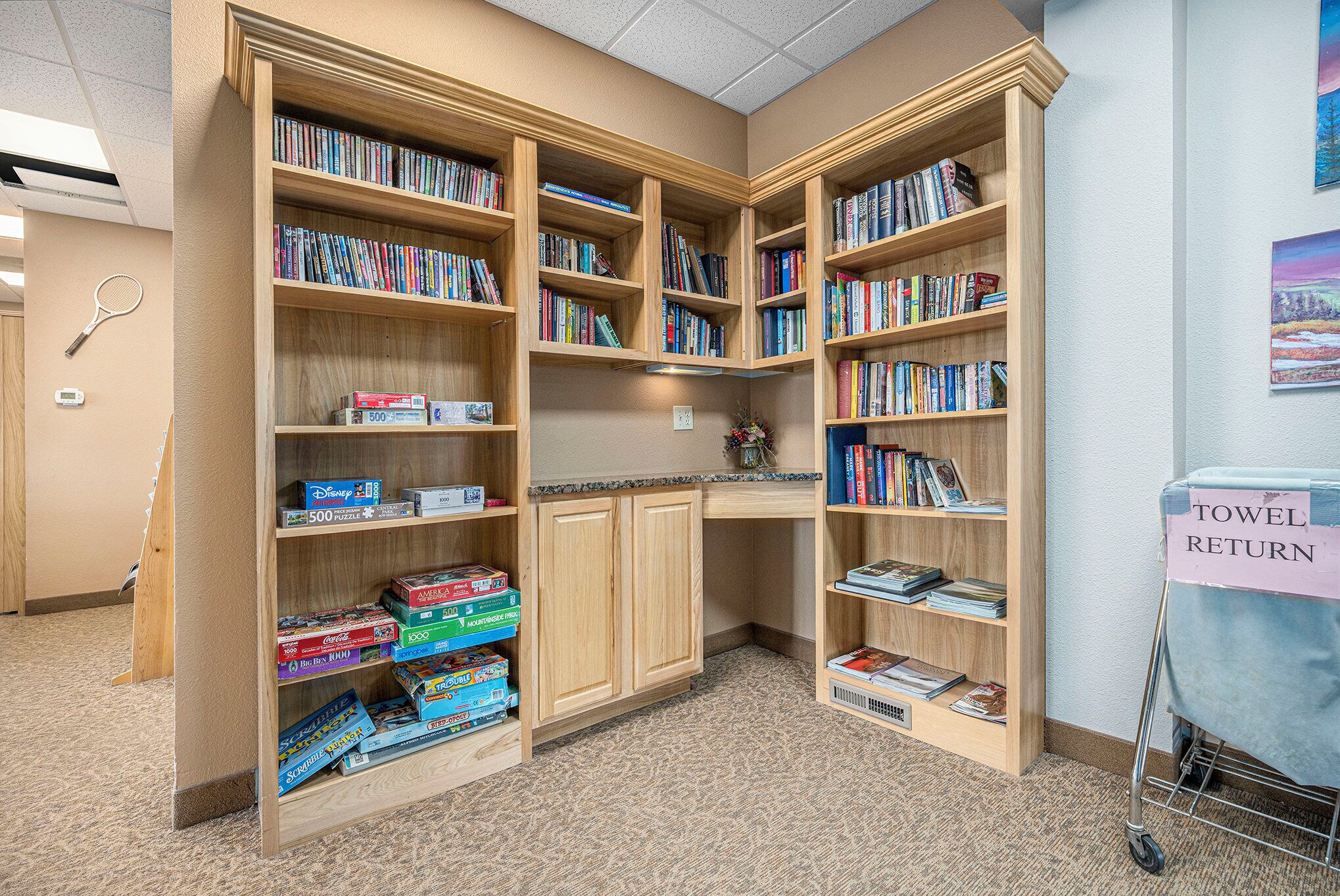 A library with wooden bookshelves filled with books and board games, a desk with a vase, and a towel return cart.