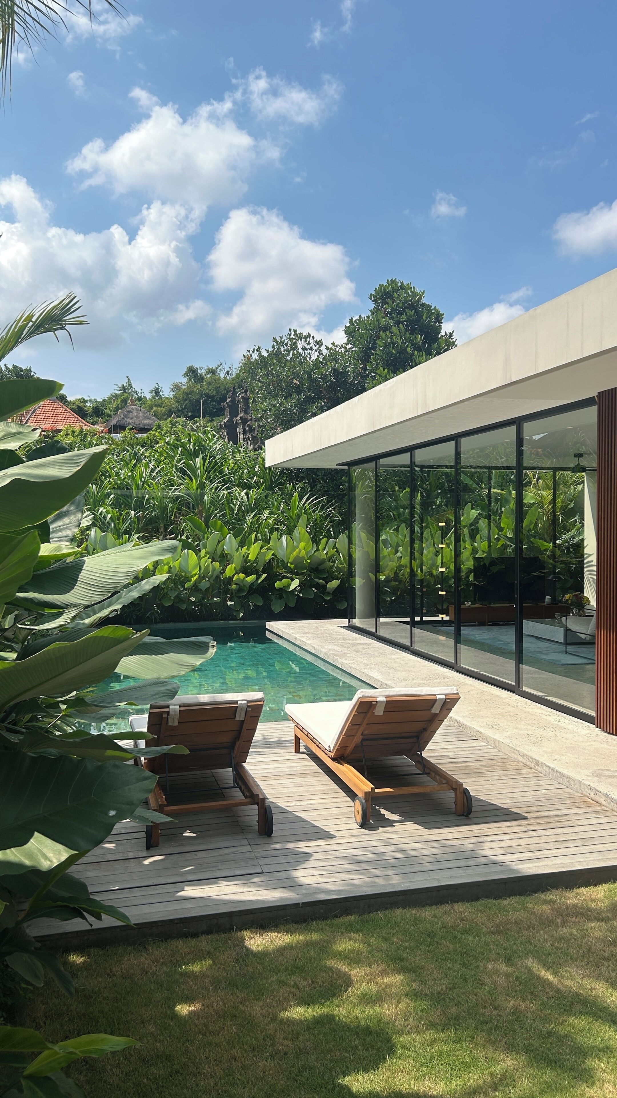 A modern outdoor pool area with two wooden lounge chairs, a glass-enclosed patio, and lush greenery.