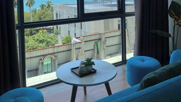 A room with a large window offering a view of a beach and mountains. There is a round table with a plant and a book, and blue upholstered furniture. - The H House Quy Nhơn (Quy Nhon)